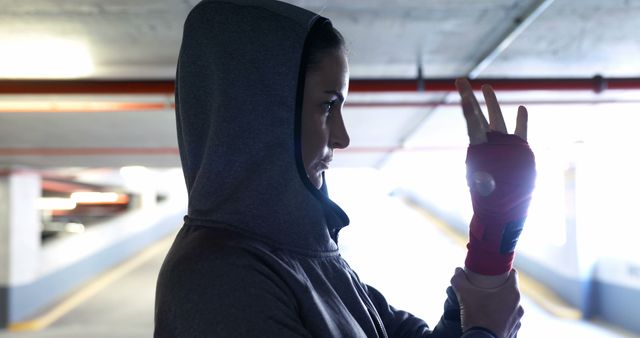 Female boxer in gray hoodie wrapping hands with red hand wraps in urban parking garage. Ideal for use in fitness magazines, motivational posters, sports ads, or urban sports fashion promotions.