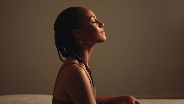 Serene woman with braids sitting on bed, eyes closed, minimalist warm profile portrait