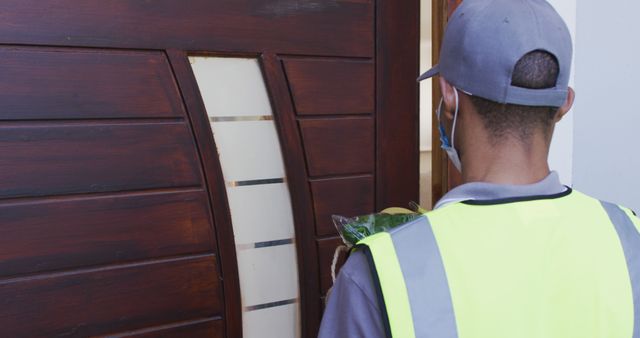 Delivery worker in high-visibility safety vest dropping off a package at the front door of a residence. Ideal for use in articles about logistics, package delivery services, online shopping, and contactless delivery options.