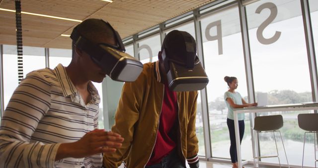 Two diverse individuals using VR headsets, engaging in immersive activities in a well-lit, modern office with large windows. Another person working on a laptop in the background suggests a blend of traditional and cutting-edge technology. Ideal for articles or marketing materials about VR, tech trends, modern workplace environments, and team collaboration.