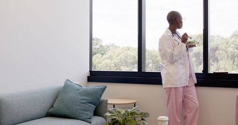 African female doctor standing by window wearing lab coat and pink scrubs during break