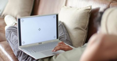 African American man reclining on leather sofa using laptop with blank screen for mockup