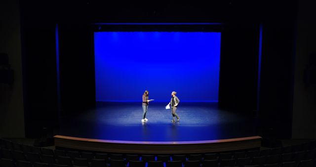 Actors practicing lines on theater stage illuminated with blue lighting, set against empty seats. Suitable for content about performing arts, theater rehearsals, behind the scenes, and drama education. Can be used in articles or designs focused on entertainment and stagecraft.