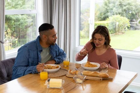 Mid-adult couple sharing homemade spaghetti at sunlit dining table with baguette and orange juice