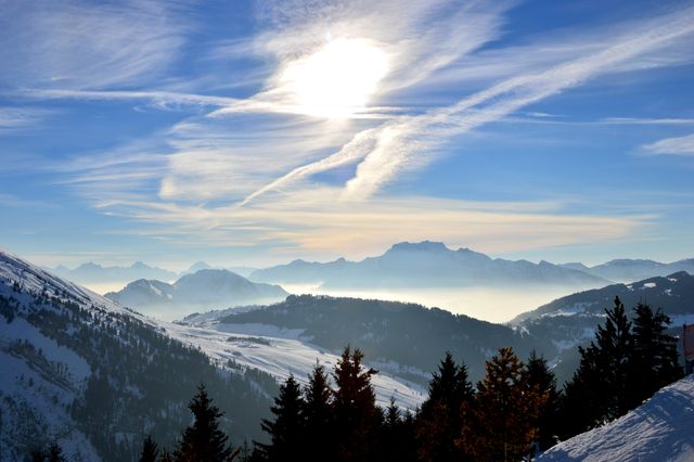 Snow-covered mountain range under a bright sun with wispy clouds spreading across the blue sky. Evergreen trees in the foreground framing the majestic view of the peaks and valleys. Image ideal for travel magazines, nature blogs, outdoor adventure brochures, environmental awareness campaigns and desktop wallpapers.