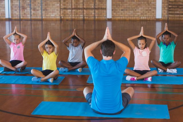 School children and their teacher practicing meditation during a yoga class in a school gym. The group is sitting on yoga mats, focusing on mindfulness and relaxation. This image can be used for educational materials, promoting physical education, wellness programs, and mindfulness activities for children.