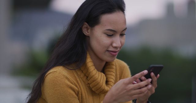 Young woman wearing yellow sweater using smartphone outdoors. Great for topics related to technology, communication, fashion, and outdoor lifestyle.