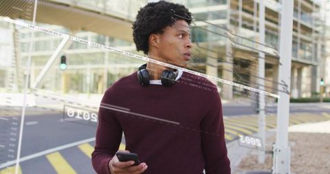 Young man standing on city sidewalk looking right holding smartphone with headphones