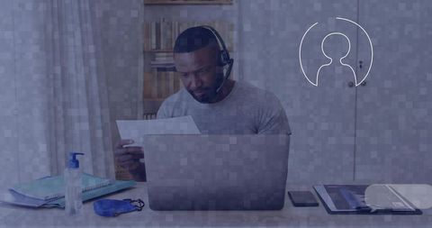 Remote worker reading document while wearing headset in home office with laptop and mask