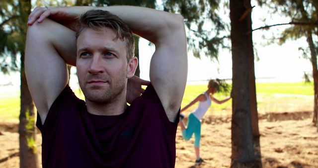 Man stretching in park with woman doing exercise in background. Perfect for fitness magazine covers, health and wellness blogs, and promotional materials for outdoor fitness programs.