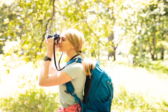 Caucasian woman with backpack taking photo in a sunlit park. Ideal for themes related to outdoor activities, nature exploration, travel, leisure, and lifestyle. Perfect for promoting camping gear, photography equipment, or travel destinations.