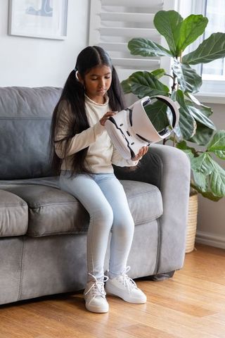 Asian school-age girl examining white vr headset while sitting on gray sofa at home