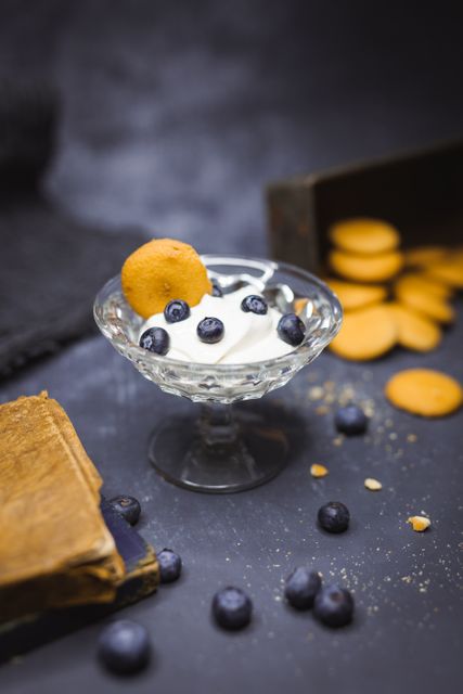 Blueberries and whipped cream served in a vintage glass dessert bowl with a biscuit on top. The dark background adds contrast, highlighting the sweetness and texture of the snack. Ideal for food-related content, blog posts, menu designs, or advertisements focusing on desserts and snacks.