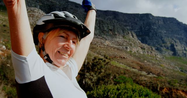 Senior woman happily raising her arms in celebration while wearing a helmet during a mountain bike ride in a scenic natural setting. Suitable for themes related to active aging, outdoor adventures, healthy living, fitness, and joyful moments in nature.