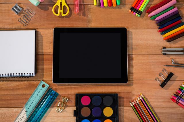 Digital tablet placed on wooden table surrounded by various school and office supplies including notepads, rulers, colored pencils, paint palette, scissors, and paper clips. Ideal for educational content, office organization tips, back-to-school promotions, or creative arts and crafts tutorials.
