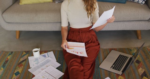 Businesswoman sitting on floor while analyzing documents, showcasing remote work setup. Ideal for content related to remote work, home office setup, work-life balance, productivity at home, and freelance or telecommuting concepts. Emphasizing casual and relaxed work atmosphere and effective document analysis at home.
