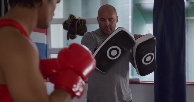 Trainer holding mitts while boxer prepares to punch in gym environment. Ideal for fitness, sports coaching, boxing training, workout routines, and combat sports promotion themes.