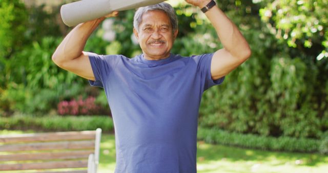 Senior man stands in a park, smiling and holding a rolled yoga mat above his head. Ideal for use in promotions about healthy lifestyle choices, fitness activities for seniors, or outdoor exercise routines.