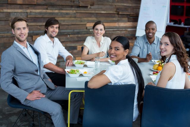 Diverse group of business professionals sitting around a table, enjoying a meal together in a modern office. They are smiling and appear engaged in conversation, suggesting a collaborative and friendly work environment. Ideal for use in articles or advertisements about corporate culture, teamwork, office life, and employee engagement.