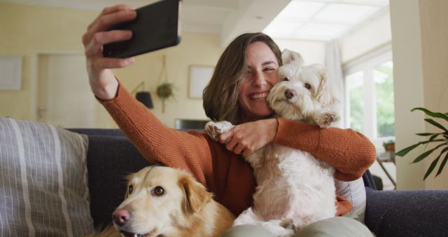 Woman holding her smartphone, capturing a selfie with two dogs on a couch in a cozy living room. The setting exudes warmth and comfort, ideal for pet lifestyle blogs, promotional content for home and pet care products, or social media campaigns celebrating everyday moments with pets.