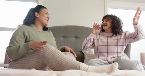 Mother and daughter laughing on bed in cozy loungewear, chatting in sunlit bedroom