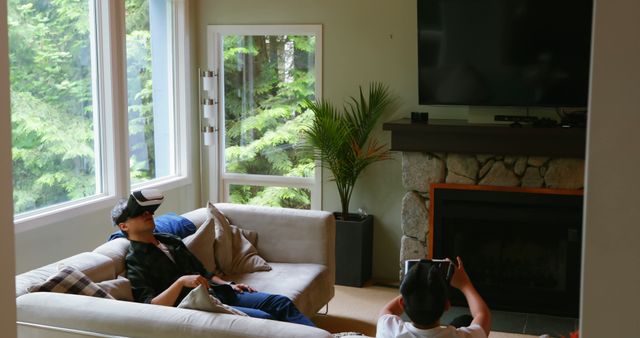 Two friends enjoying leisure time in a modern living room where they are relaxing on a sofa and seated on the floor wearing virtual reality headsets. The living room features large windows with a view of lush green trees outside, a fireplace, and a potted plant. Can be used for topics on technology in everyday life, leisure activities, modern living spaces, and friendships.