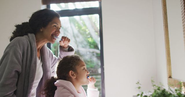 Mother and daughter are engaging in morning brushing routine together, smiling in front of bathroom mirror. Perfect for depicting family health care, daily hygiene practices, and parent-child bonding. Ideal for use in oral care advertisements, parenting blogs, and family health guides.