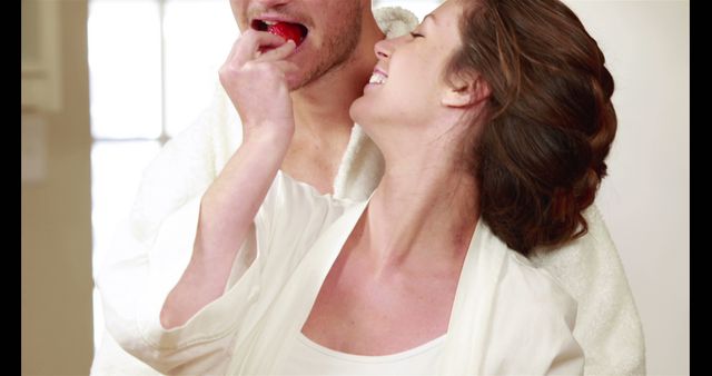 A Caucasian woman is feeding a strawberry to a man, both appear joyful and are wearing white bathrobes, with copy space. Their playful interaction suggests a romantic or intimate moment, in a relaxed, domestic setting.