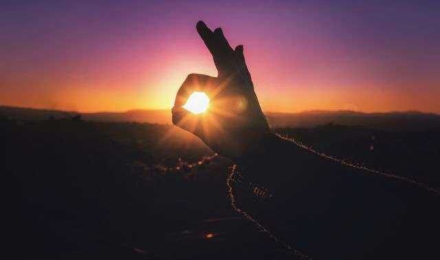 Hand is forming OK sign against a vibrant sunset sky. Orange and purple hues create serene and calm atmosphere. Can be used for themes relating to communication, positivity, evaluation, or nature. Suitable for websites, blogs, social media posts, inspirational content, and mental wellness campaigns.