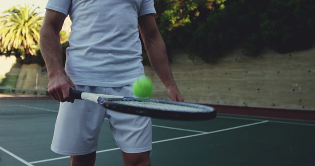 Young adult practicing tennis on an outdoor court, bouncing a tennis ball on the racket. Excellent for content on sports training, active lifestyles, outdoor activities, tennis skills, and fitness routines.