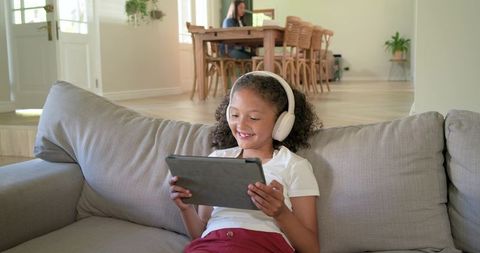 Mother working while daughter using tablet on sofa in bright open-plan living space
