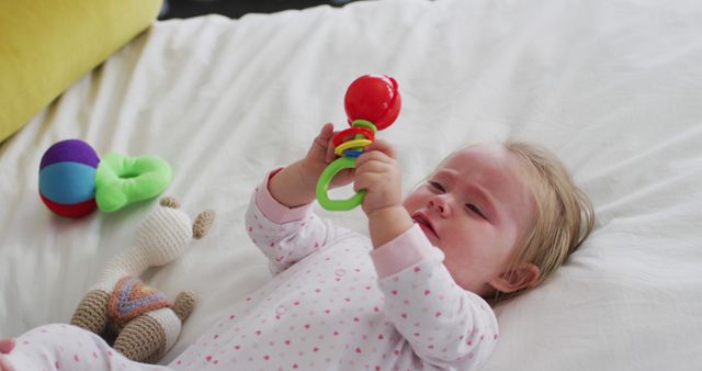 A baby lies on a bed while holding and examining a colorful rattle. Clothes and toys can also be seen, suggesting a playful and joyful atmosphere. Suitable for promoting baby products, early learning tools, parenting blogs, and related content that deals with infant development and care.
