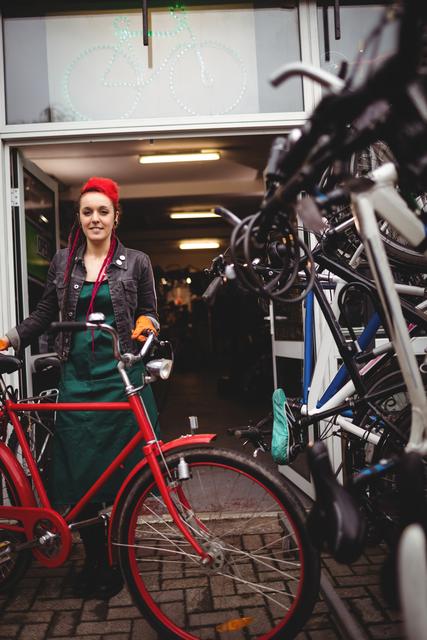 Smiling female mechanic standing with a red bicycle in a workshop. She is wearing an apron and appears ready to repair or service the bike. This image can be used for promoting bicycle repair services, cycling workshops, or professional mechanic services. It is also suitable for articles or advertisements related to cycling, transportation, and maintenance.