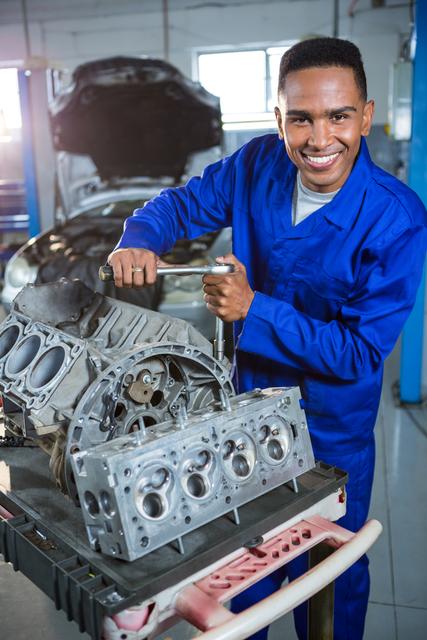 Mechanic in blue overalls smiling while repairing a car engine in a well-lit garage. Ideal for use in automotive service advertisements, mechanical engineering articles, and professional training materials.