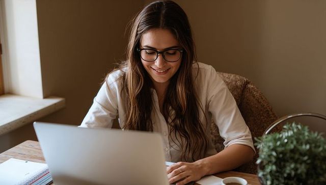 Smiling woman typing on laptop at sunlit home office desk with glasses, coffee, plant