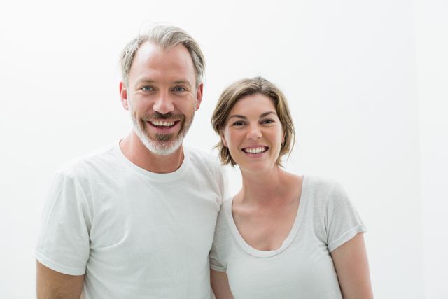 Smiling couple standing close together in bedroom, both wearing white t-shirts. Ideal for use in lifestyle blogs, relationship advice articles, home decor websites, and advertisements promoting casual clothing or home products.