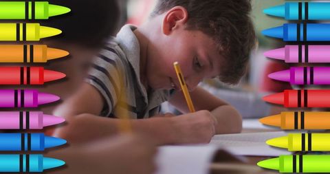 Focused schoolboy writing surrounded by colorful crayons