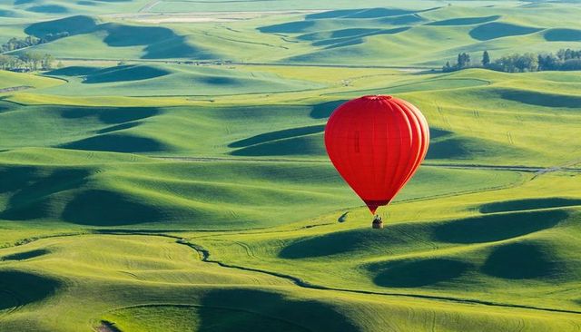 Red hot air balloon drifting over rolling green hills at sunrise scenic aerial landscape