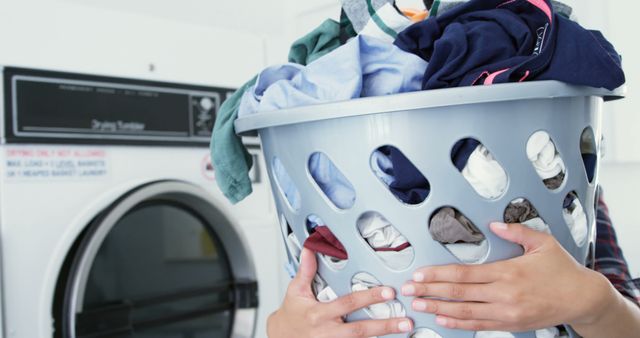 Hands of biracial woman carrying full laundry basket of clothes at laundromat, copy space. Laundry, washing machine, tumble dryer, domestic chores and local business, unaltered.