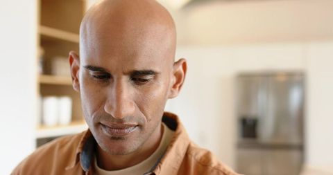 Contemplative bald mature man standing in bright modern kitchen looking down in thought