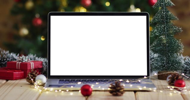 Laptop with blank screen surrounded by Christmas decorations, including a miniature Christmas tree, pine cones, and wrapped presents. Christmas lights add a festive touch to the wooden surface. Perfect for holiday-themed advertisements, festive promotions, or illustrating a holiday workspace.