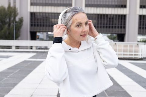 Mature woman adjusting wireless headphones on urban rooftop plaza wearing white hoodie