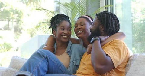 Smiling family trio cuddling on sunlit sofa sharing warm home moment