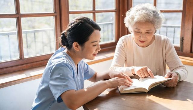 Caregiver guiding senior woman reading together at sunlit wooden table for compassionate care