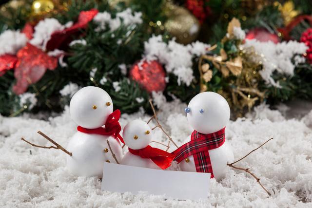 Three snowmen with red and plaid scarves are surrounded by fake snow and festive decorations. Ideal for holiday greeting cards, Christmas advertisements, winter-themed promotions, and festive decor inspiration.