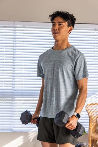 Asian man lifting hex dumbbells in sunlit living room wearing athletic tee and smartwatch