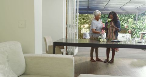 Senior woman and African-American woman arranging white rose bouquet on modern dining table