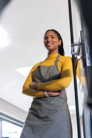 Smiling barista standing at storefront wearing mustard top and gray apron