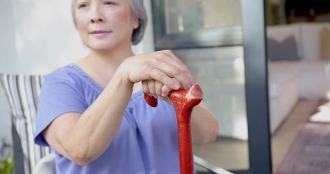 Serene senior Asian woman sitting on porch holding red cane wearing lavender top