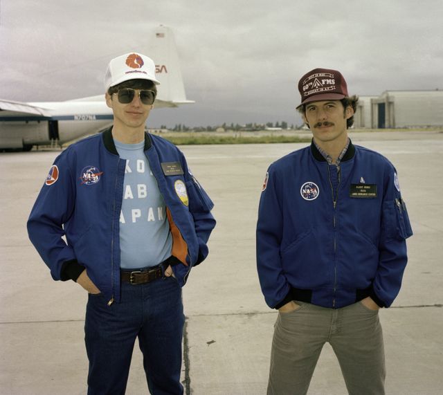 Two NASA scientists standing outdoors at Ames Research Center, California in casual clothing, preparing for departure to Australia. They are wearing NASA jackets and caps with aircraft visible in the background. Potential use in articles or materials related to NASA missions, aviation, space science, and international research collaborations.
